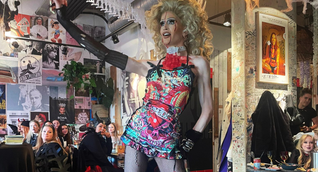 Dragqueen in kleurrijke jurk treedt op in een zaal met publiek en kunst aan de muren