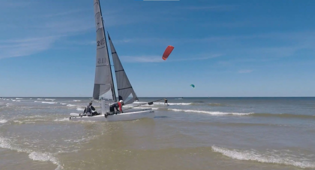 Catamaran met opgestoken zeilen vaart de zee op, met kitesurfers verderop in beeld onder blauwe lucht