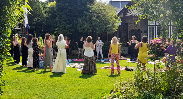 Groep mensen doet mee aan een mindful buitenritueel of workshop in een groene tuin bij zonnig weer.