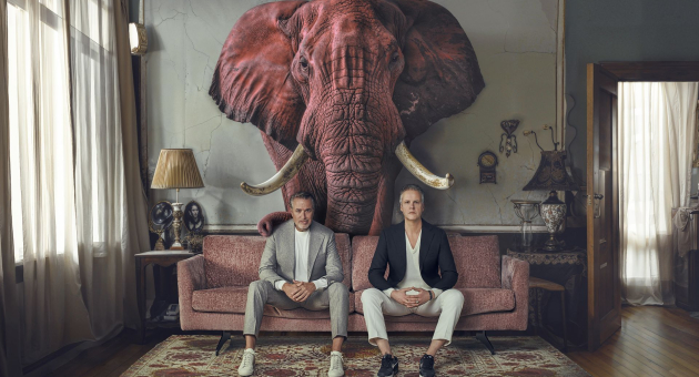 Veldhuis en kemper zitten op een bank in een woonkamer met een roze olifant achter zich. 