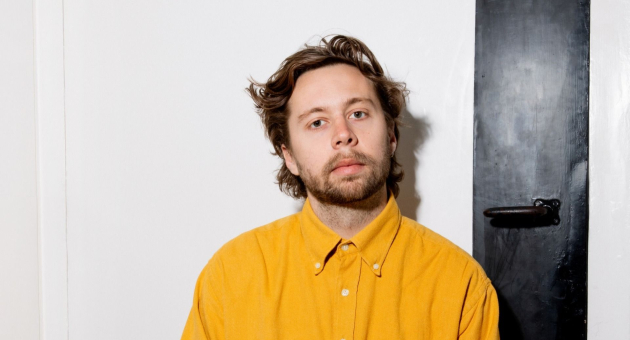 Een jonge man met halflang haar en een baard kijkt neutraal in de camera. Hij draagt een felgele overhemd en zit tegen een witte muur met een zwarte deur of paneel aan de rechterkant.