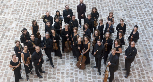en orkest poseert in een halve cirkel op een binnenplein met een stenen vloer. De muzikanten dragen zwarte kleding en houden hun instrumenten vast, terwijl één persoon in een donkerrood pak centraal achter hen staat. Ze kijken omhoog naar de camera.