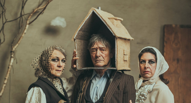 Drie mensen in theatrale kostuums poseren met serieuze gezichten; de persoon in het midden draagt een huis als hoofddeksel.