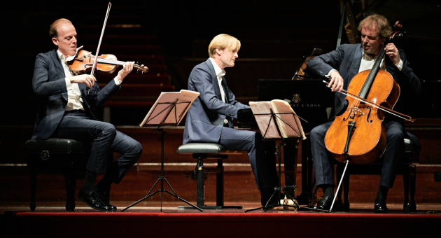 Drie muzikanten voeren een klassiek pianotrio uit op een concertpodium, met viool, piano en cello.