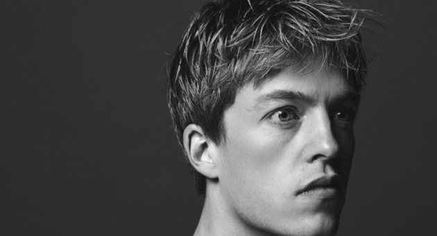 Zwart-wit portret van een jonge man met warrig kort haar, die serieus en geconcentreerd naar rechts kijkt tegen een donkere achtergrond.