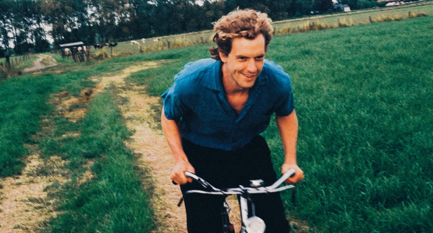 De foto toont een jonge man met een krullend kapsel die lachend op een fiets rijdt over een smal, onverhard pad door een groen landschap. 