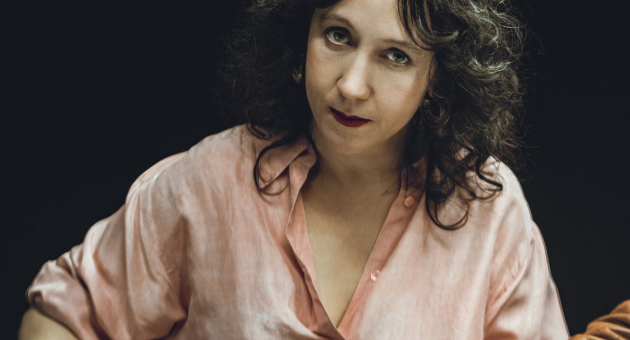 Een vrouw met donker krullend haar en een lichte, openvallende blouse kijkt serieus en recht in de camera, tegen een donkere achtergrond.