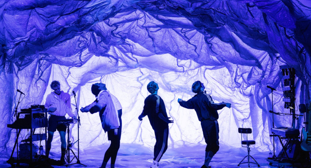 Vier mensen dansen onder een kunstzinnig verlicht decor dat lijkt op een grot van paarse doeken, met een muzikant links die live muziek maakt.