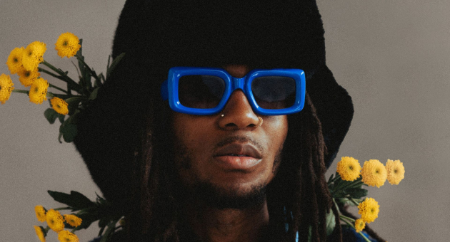 Persoon met zwarte hoed en felblauwe zonnebril, omringd door gele bloemen tegen neutrale achtergrond.