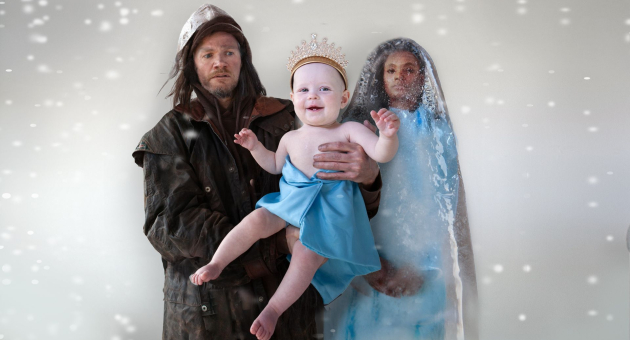 Man en vrouw in winterse kleding houden baby met blauwe doek en kroon vast in vallende sneeuw.