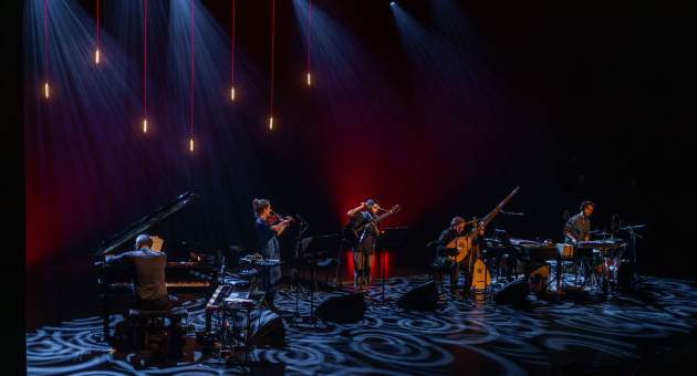 Muzikanten op een donker podium met blauwe en rode verlichting tijdens een liveconcert, met piano, percussie en blaasinstrumenten