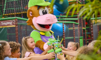 Een vrolijke mascotte in de vorm van een aap deelt bekers uit aan lachende kinderen in een kleurrijke indoor speeltuin.