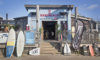 Ingang Hart Beach surfschool bestaande uit lichtblauwe houten panelen en rood logo. Er staan surfplanken op een rij voor de deur naast de trap.