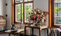 Historisch interieur met houten meubels, droogbloemen op een tafel en uitzicht op grachtenpand door glas-in-loodramen