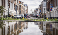 Een drukke straat in Den Haag met voetgangers en auto's, weerspiegeld in een plas water.