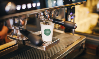 Kartonnen beker met logo van Dudok onder een professioneel koffieapparaat. 