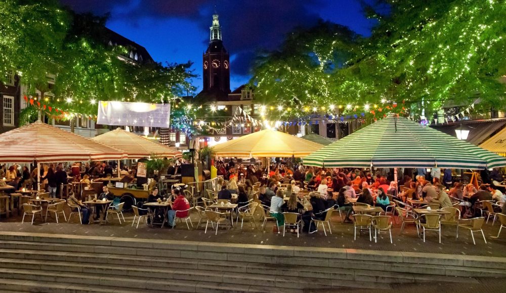 Het drukke terras van de Grote Markt in Den Haag in de avond. Mensen zitten onder parasols op het sfeervol verlichtte terras.
