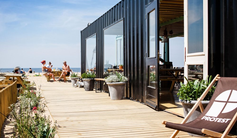 Mensen lekker genietend van de zon op het zij-terras van The Shore op Scheveningen. Deze duurzame strandtent bevindt zich in een container.