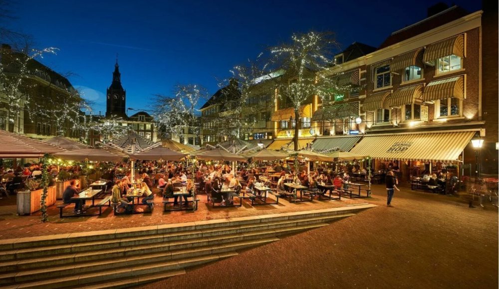 De terrassen op de Grote markt gezellig verlicht in de avond. Op de achtergrond het silhouet van de Grote Kerk.