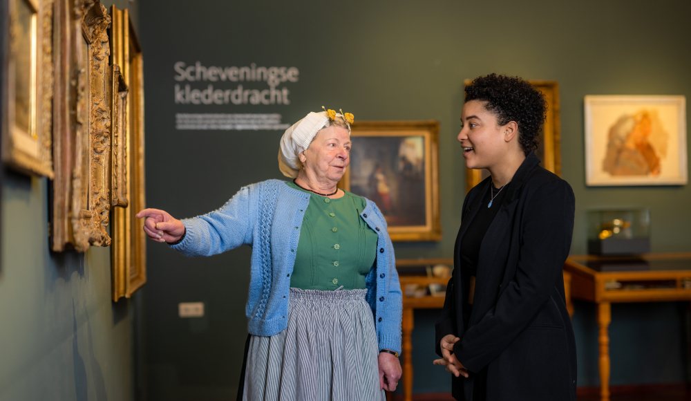 Vrouw in klederdracht wijst iets aan op een schilderij in Muzee Scheveningen