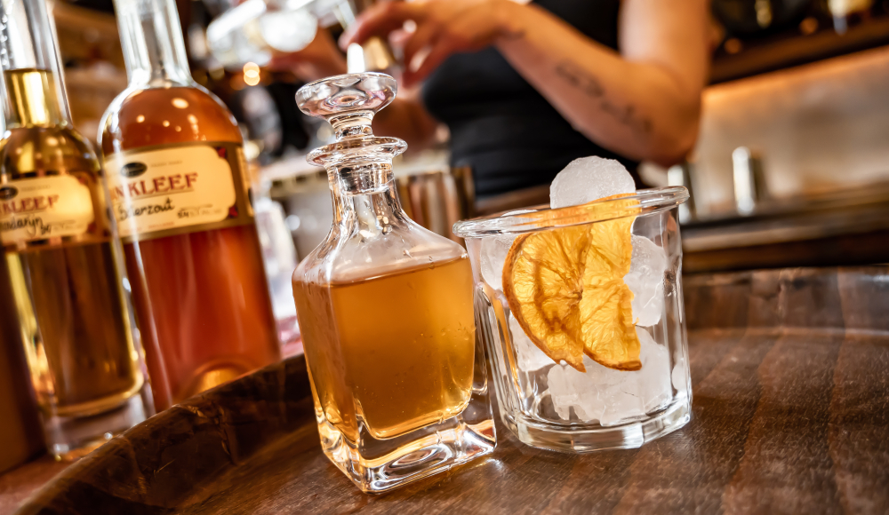 Cocktail Old Louis in klassiek glazen karaf met stop en glas met gedroogde sinaasappelschijfjes en ijsklontjes op dienblad op counter met serveerster vaag op de achtergrond.