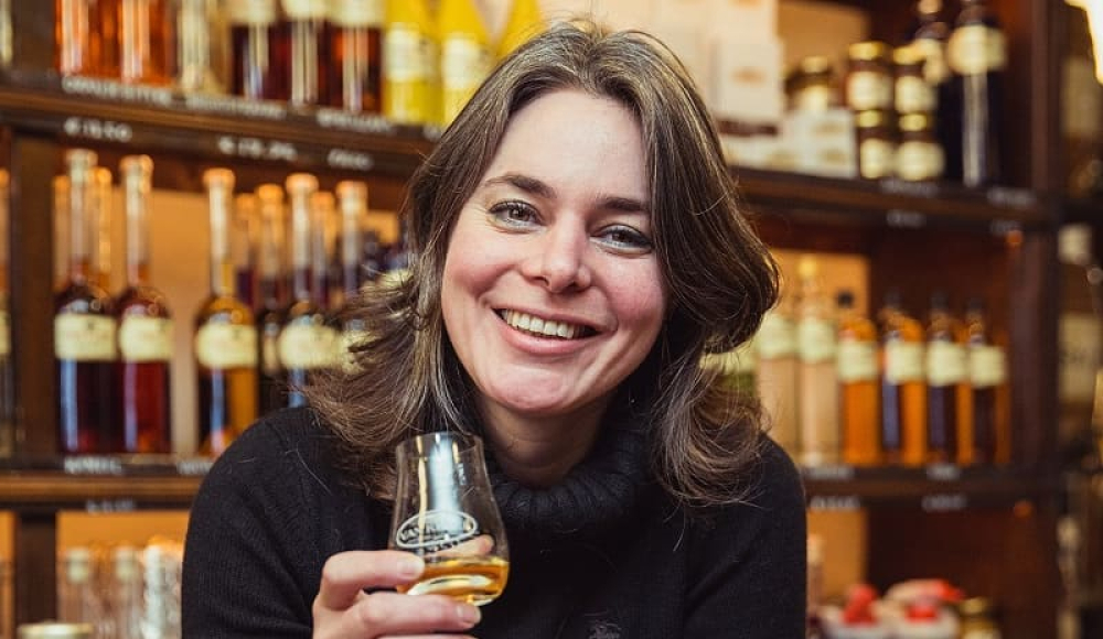 Fleur Kruyt met glas Van Kleef likeur aan de counter voor een schap flessen