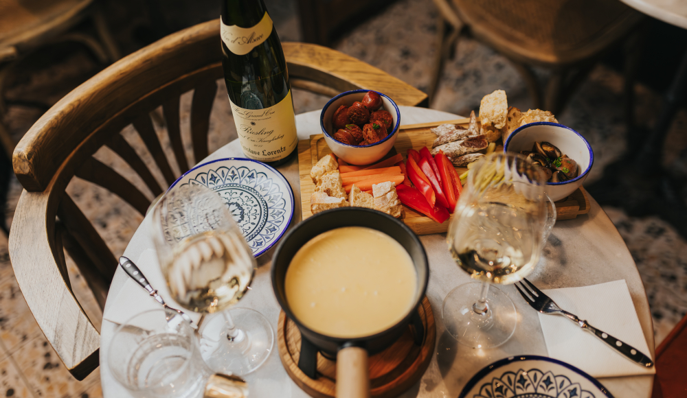 Gedekte tafel bij Vinostra met een kaasfondue pannetje met verschillende garnituren ernaast
