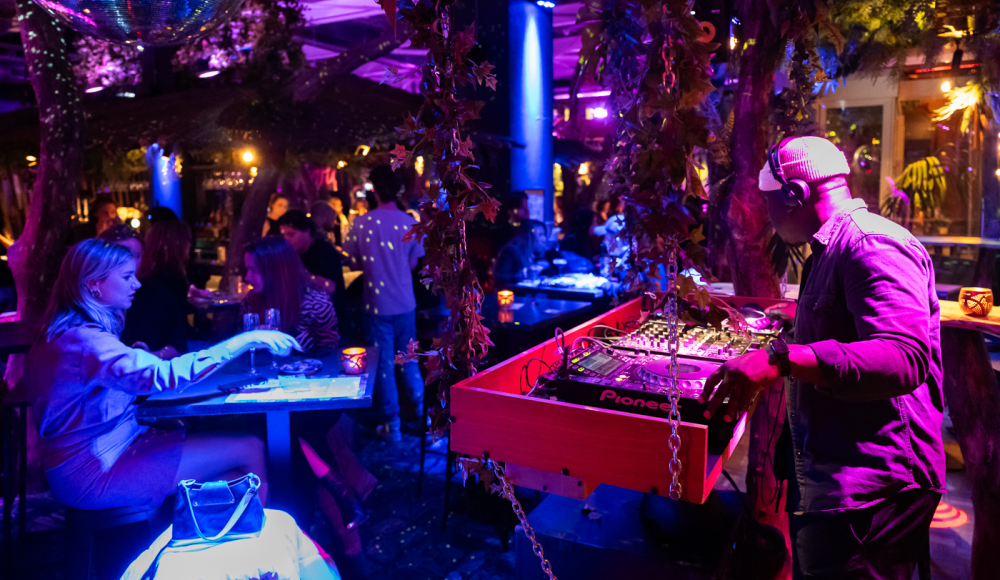 De foto toont een sfeervolle uitgaanssetting met een DJ die live muziek draait op een mengpaneel, omringd door kleurrijke paarse en blauwe verlichting. De locatie heeft een exotische en intieme ambiance, met groene planten en decoratieve elementen zoals een discobal aan het plafond. 