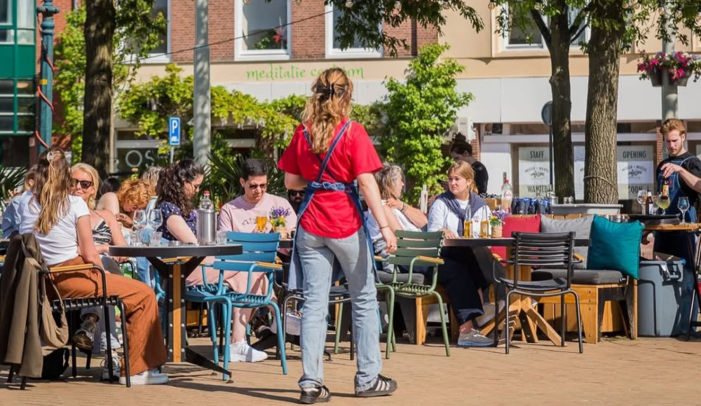 Een levendig terras op een zonnige dag in Den Haag. Mensen genieten van een drankje aan kleurrijke tafels onder de groene bomen, terwijl een serveerster in een rode blouse langsloopt om bestellingen op te nemen. Een levendig terras op een zonnige dag in Den Haag. Mensen genieten van een drankje aan kleurrijke tafels onder de groene bomen, terwijl een serveerster in een rode blouse langsloopt om bestellingen op te nemen.