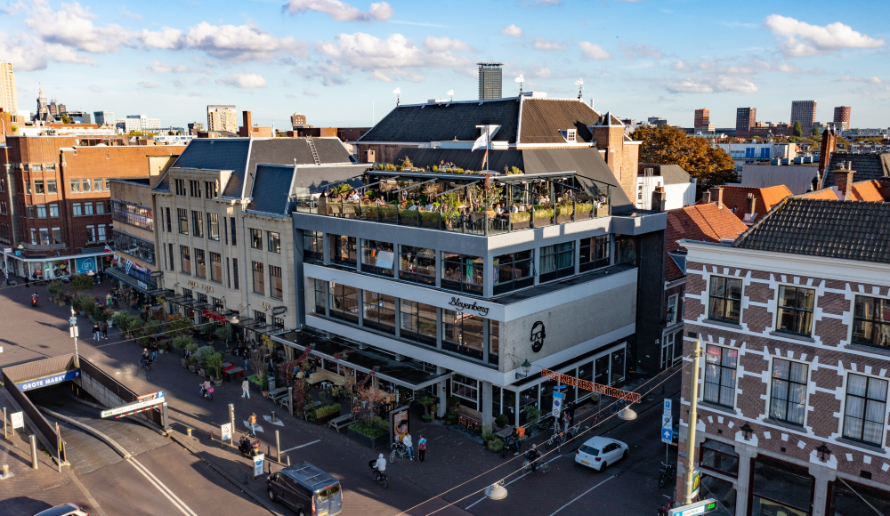 De foto toont een levendig stadsbeeld met het opvallende pand van Bleyenberg in het midden. 