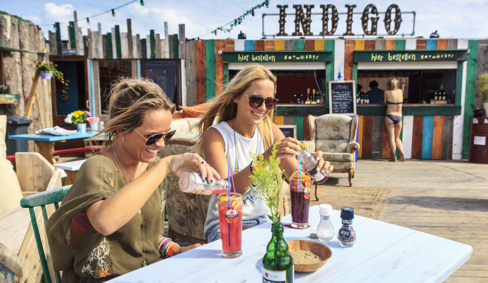 Twee dames genieten van een drankje op het strand bij Beachclub Indigo Twee dames genieten van een drankje op het strand bij Beachclub Indigo
