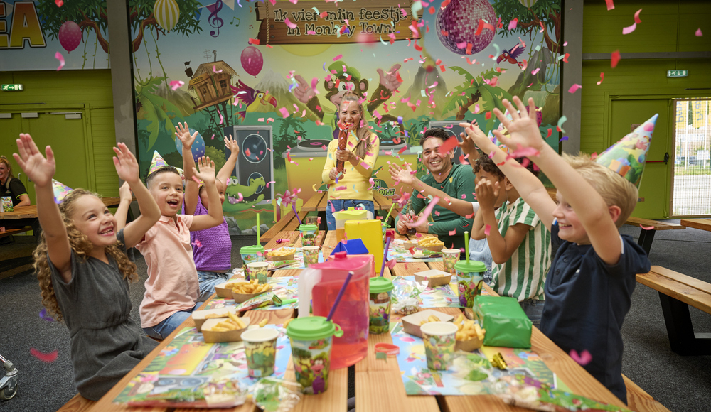Kinderen vieren een vrolijk verjaardagsfeest bij Monkey Town, zittend aan een kleurrijk versierde tafel terwijl confetti door de lucht vliegt.