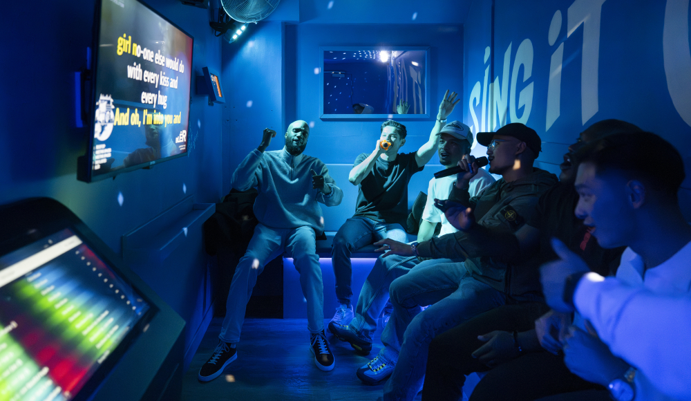 Een groep mannen zit in een blauw verlichte karaokekamer en zingt mee met het scherm.