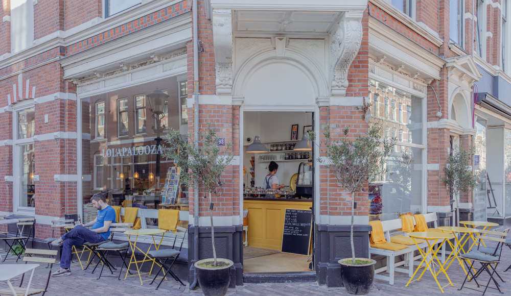 Gezellige koffiebar op een hoekpand in de Weimarstraat met geel meubilair op het terras en mensen die buiten zitten te genieten.