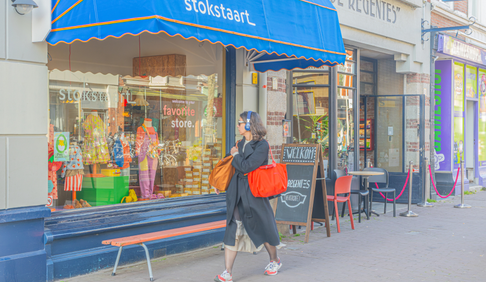 Vrouw loopt langs kleurrijke etalage van speelgoedwinkel Stokstaart in de Weimarstraat, Den Haag, met een grote oranje tas.