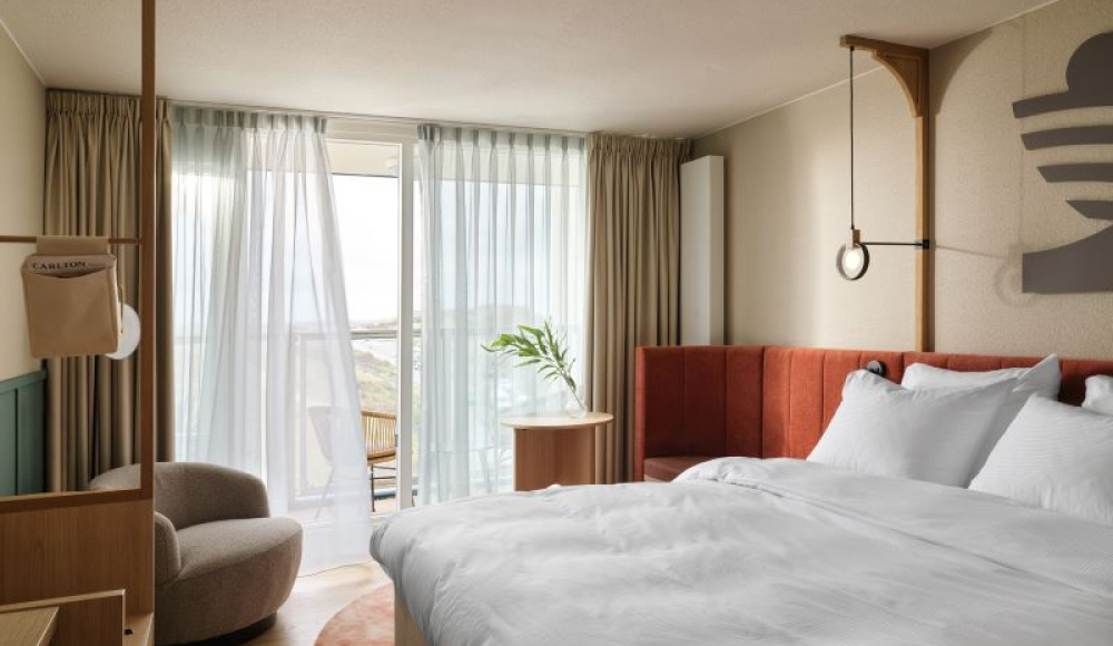 Licht en modern hotelkamerinterieur met een groot wit bed, warme houten accenten, een ronde fauteuil en uitzicht op een zonnig balkon met gordijnen die zachtjes waaien. Licht en modern hotelkamerinterieur met een groot wit bed, warme houten accenten, een ronde fauteuil en uitzicht op een zonnig balkon met gordijnen die zachtjes waaien.