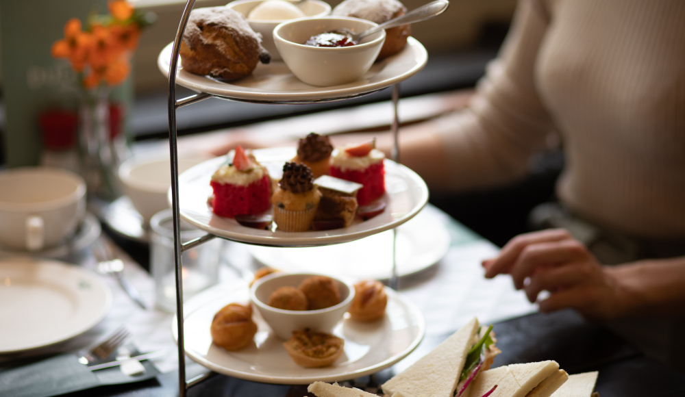 High tea etagère met scones, gebakjes en hartige hapjes op tafel, met sandwiches op een plank en een persoon High tea etagère met scones, gebakjes en hartige hapjes op tafel, met sandwiches op een plank en een persoon