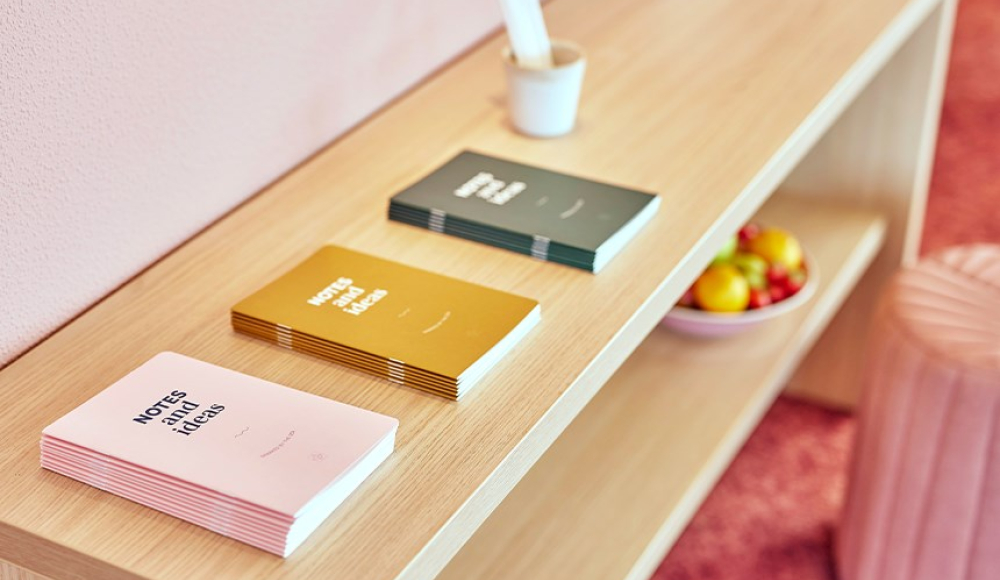 Close-up van een houten plank met kleurrijke notitieboekjes, een kopje met pennen en een schaal met fruit in een modern, zachtroze interieur.