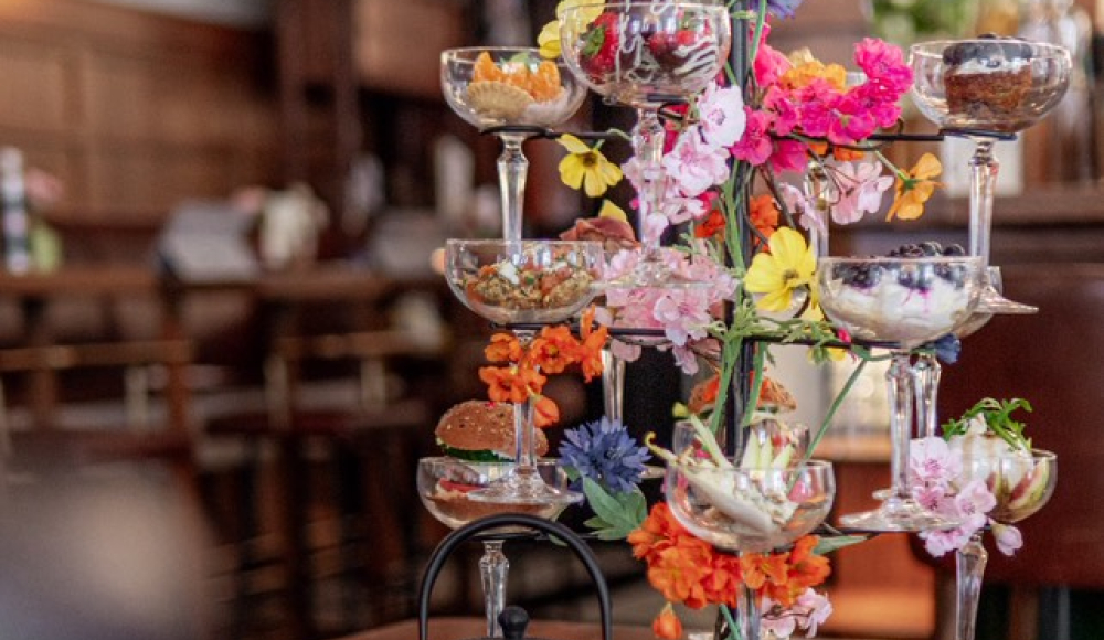 Gedekte tafel met een opvallende en kleurrijke high tea etagère in glazen, versierd met bloemen, en een zwarte theepot in een sfeervol restaurantinterieur.