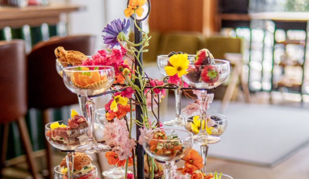 Kleurrijke en creatieve high tea geserveerd in glazen op een etagère, versierd met bloemen, met een theepot en servies op tafel.