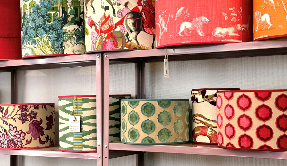 Kleurrijke handgemaakte lampenkappen met diverse patronen, zoals dierenprints, botanische motieven en grafische designs, uitgestald op metalen planken in een winkel. Kleurrijke handgemaakte lampenkappen met diverse patronen, zoals dierenprints, botanische motieven en grafische designs, uitgestald op metalen planken in een winkel.