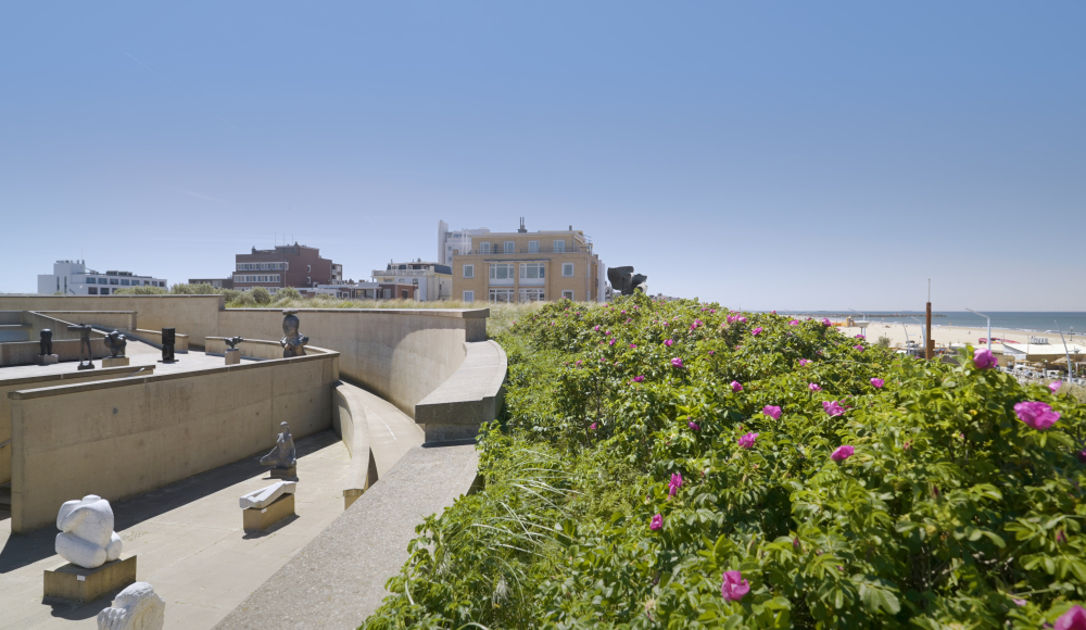 Uitzicht op het buitenterrein van Museum Beelden aan Zee met beelden, bloeiende struiken en op de achtergrond het strand en de zee van Scheveningen. Uitzicht op het buitenterrein van Museum Beelden aan Zee met beelden, bloeiende struiken en op de achtergrond het strand en de zee van Scheveningen.