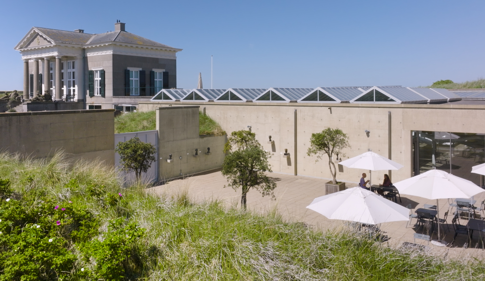 Binnenplaats van Museum Beelden aan Zee met terrastafels en witte parasols, omringd door duingras en met uitzicht op het historische Paviljoen Von Wied.