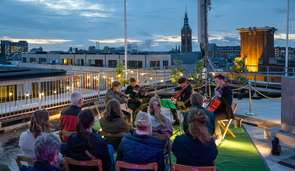 Openluchtconcert met strijkkwartet op een dakterras van de Bijenkorf in Den Haag, met uitzicht op de skyline en de Grote Kerk Openluchtconcert met strijkkwartet op een dakterras van de Bijenkorf in Den Haag, met uitzicht op de skyline en de Grote Kerk