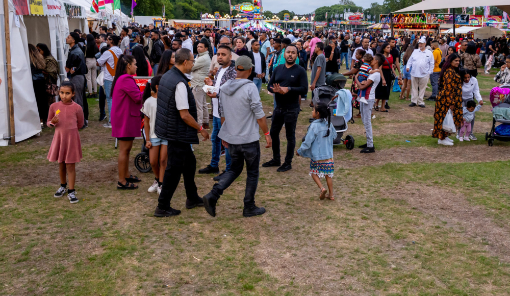 Bezoekers wandelen over een druk bezocht festivalterrein in het Zuiderpark, met kraampjes, eettentjes en kleurrijke vlaggen. Bezoekers wandelen over een druk bezocht festivalterrein in het Zuiderpark, met kraampjes, eettentjes en kleurrijke vlaggen.