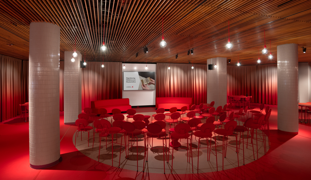 Zaalopstelling met rode stoelen in halve cirkel, podium met scherm, houten plafond en pilaren in modern interieur
