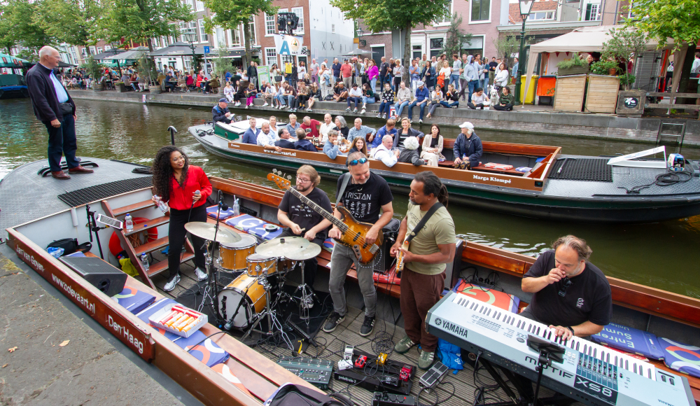 Band speelt op een boot in de gracht, publiek kijkt toe vanaf kade en andere boten in een gezellige binnenstad