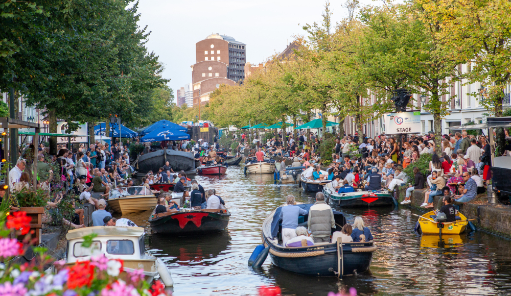 Drukke gracht vol boten met mensen, publiek zit langs de kade tussen bomen en terrassen in zomerse sfeer