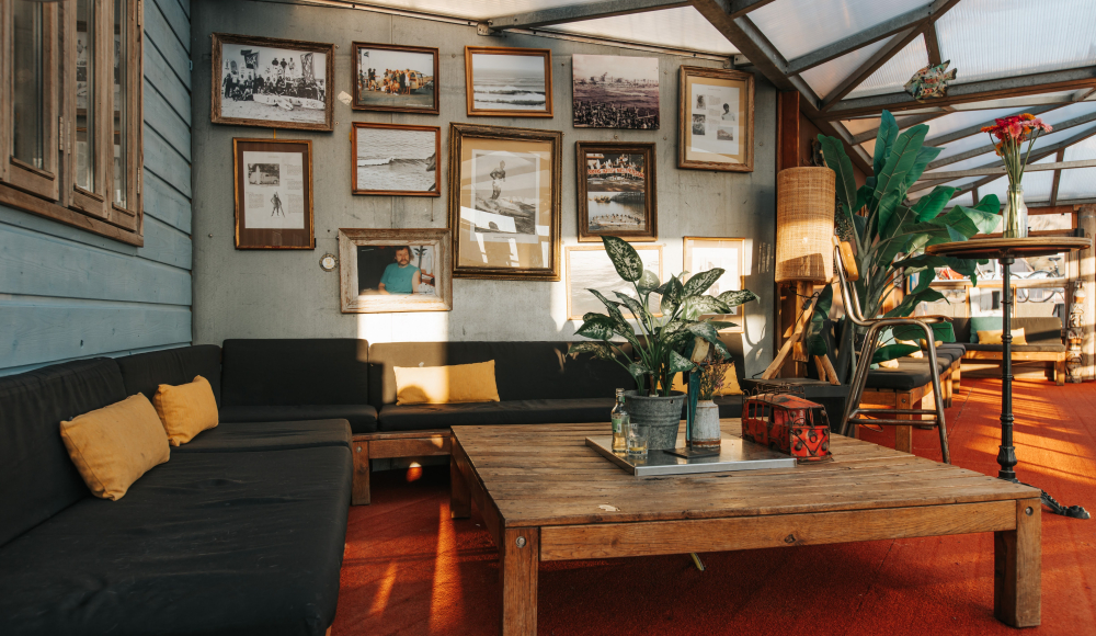 Hartbeach sfeervolle interieur impressie met ingelijst surffoto's aan de wand, een grote bruine hoekbank met gele kussens, planten en een houten tafeltje met kaarsplateau Foto: Gabriel Zajicek Hartbeach sfeervolle interieur impressie met ingelijst surffoto's aan de wand, een grote bruine hoekbank met gele kussens, planten en een houten tafeltje met kaarsplateau Foto: Gabriel Zajicek