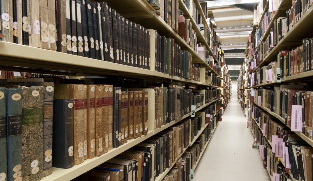 Smalle gang tussen hoge archiefrekken vol oude boeken en gebonden tijdschriften in depotomgeving